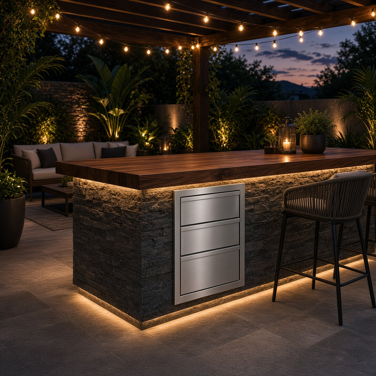 Outdoor kitchen island with a wood-look countertop, stone-look lower cabinet panel, and a stainless steel three-drawer front, shown under a pergola at dusk with string lights and patio seating in the background.